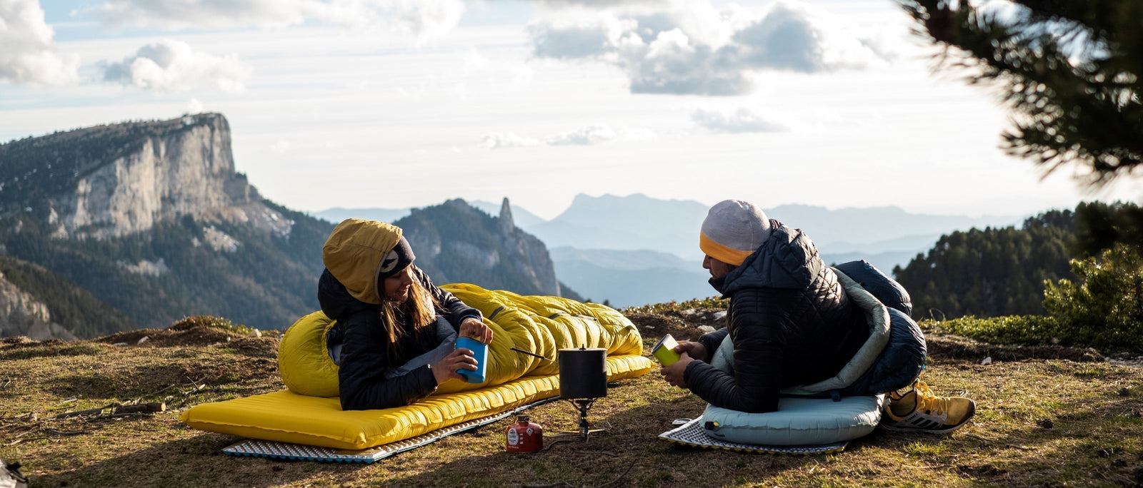 A man and a woman lying on sleeping mats and bags on an open hillside, with a dramatic mountain cliff and wide alpine valley behind them, sharing a hot drink from a camping stove.