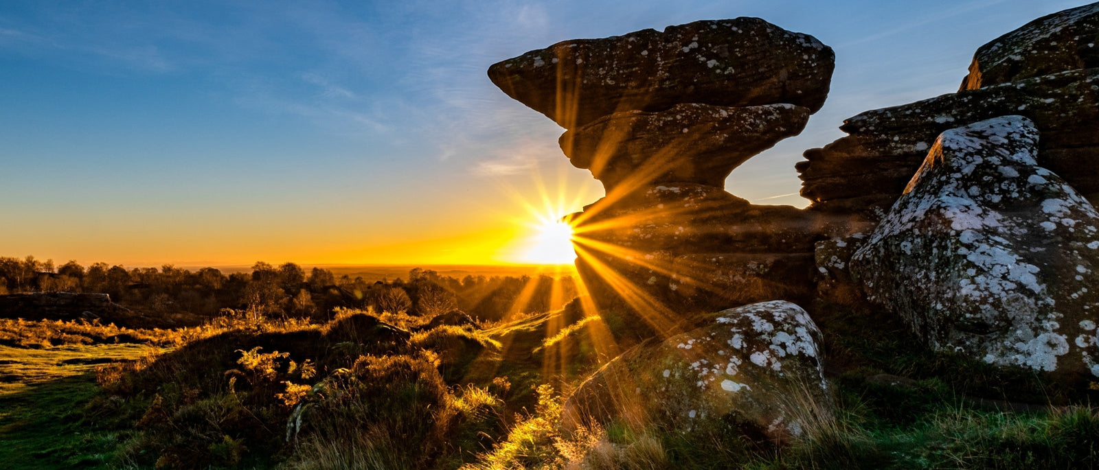 Sunrise at Brimham Rocks with sun's rays bursting through gap in precariously balanced millstone grit rock formation, with weathered stone, golden heather and distant woodland silhouetted against blue and orange sky.