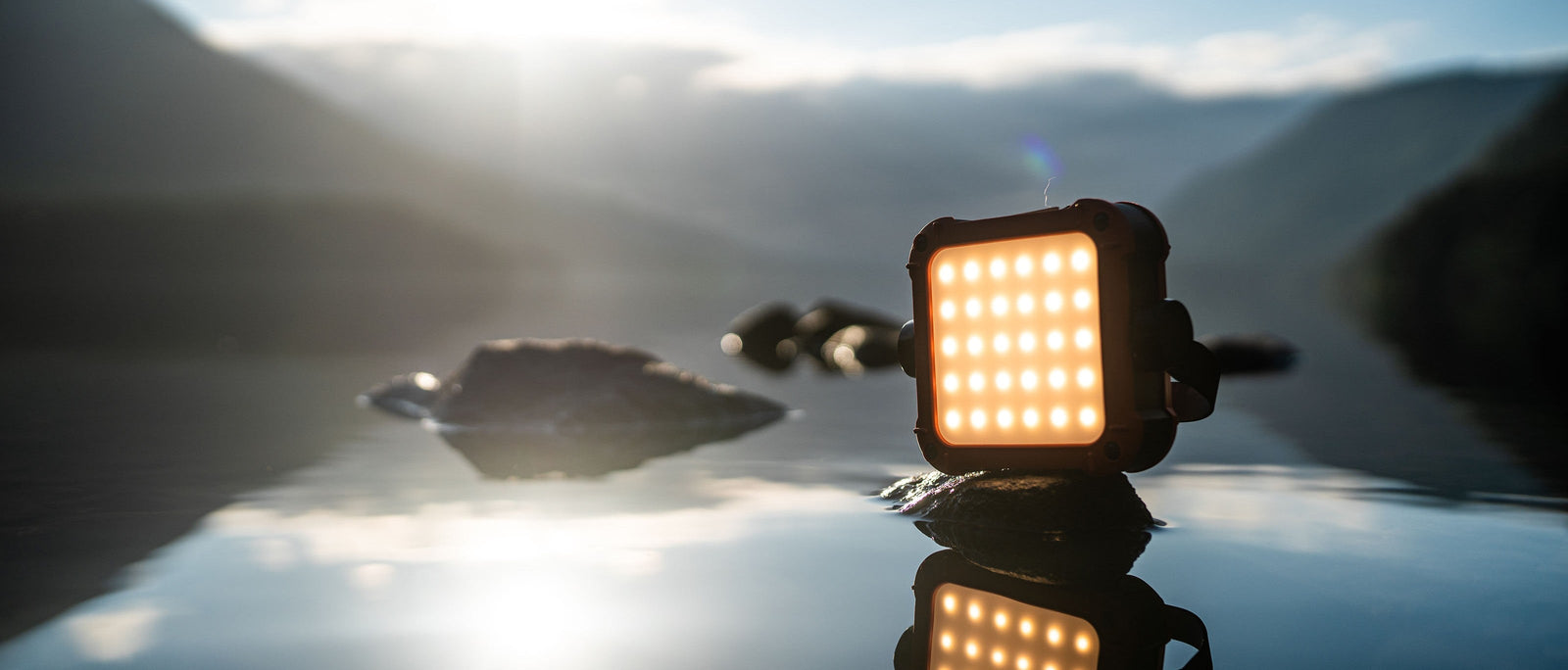 Compact Claymore Ultra LED lantern balanced on rock reflecting in still mountain lake water at dawn.