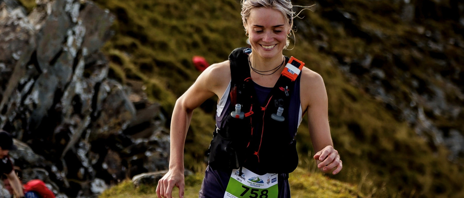 Trail runner Sophie Ranson with a running vest stuffed full of soft flasks, smiling as she runs uphill on a rocky mountain trail during the Ring of Steall Skyrace.