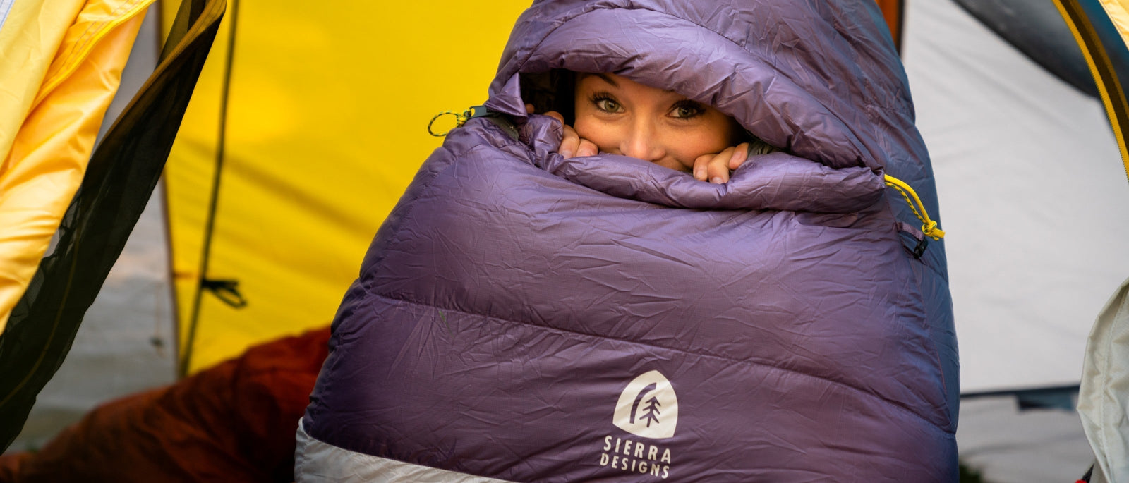 A woman cocooned inside a purple mummy sleeping bag with only her eyes and nose visible, sitting in a tent doorway