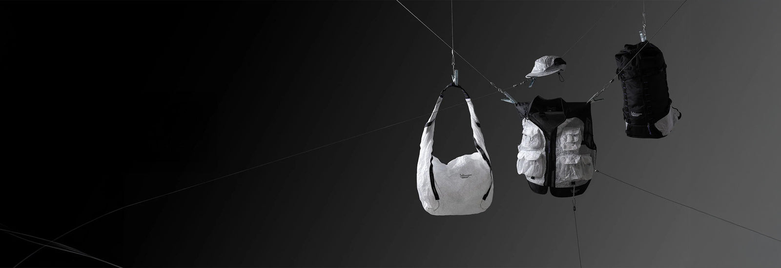Outdoor gear suspended on ropes against a dark background, including a white and black shoulder bag, fishing vest, cap