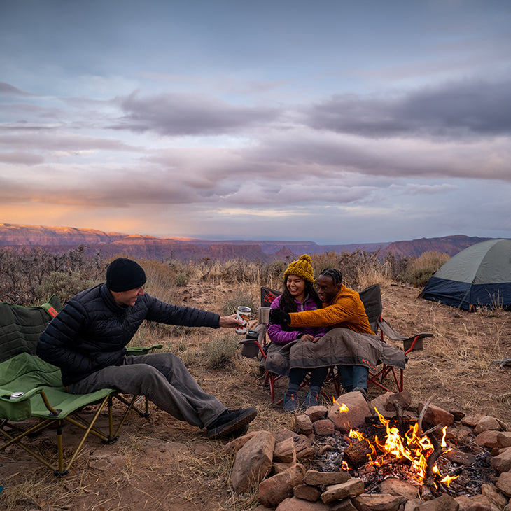 Three campers toasting drinks by a campfire with tents and mountains at sunset.