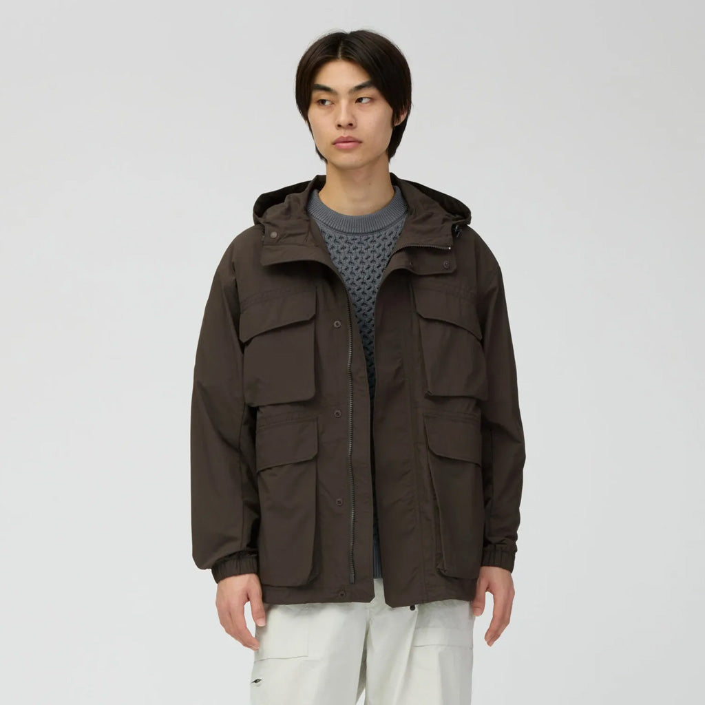 Snow Peak men's brown hooded jacket with multiple chest pockets, worn with grey knit and cream trousers.