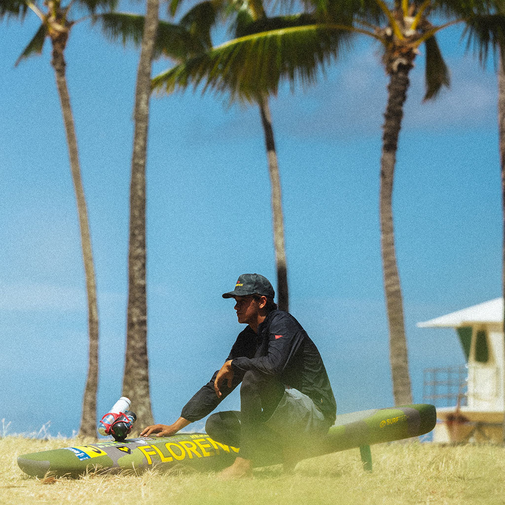A person preparing a surfboard on a tropical beach with palm trees and clear blue sky.
