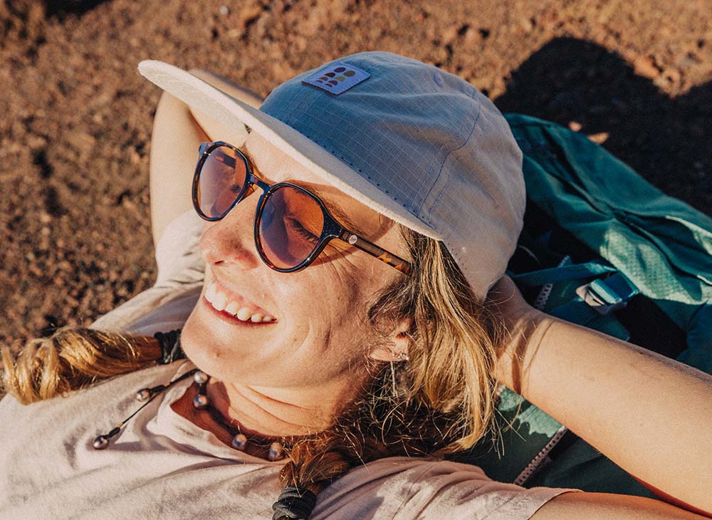 A person wearing Sunski sunglasses and a blue cap relaxes on sandy ground in bright sunshine.