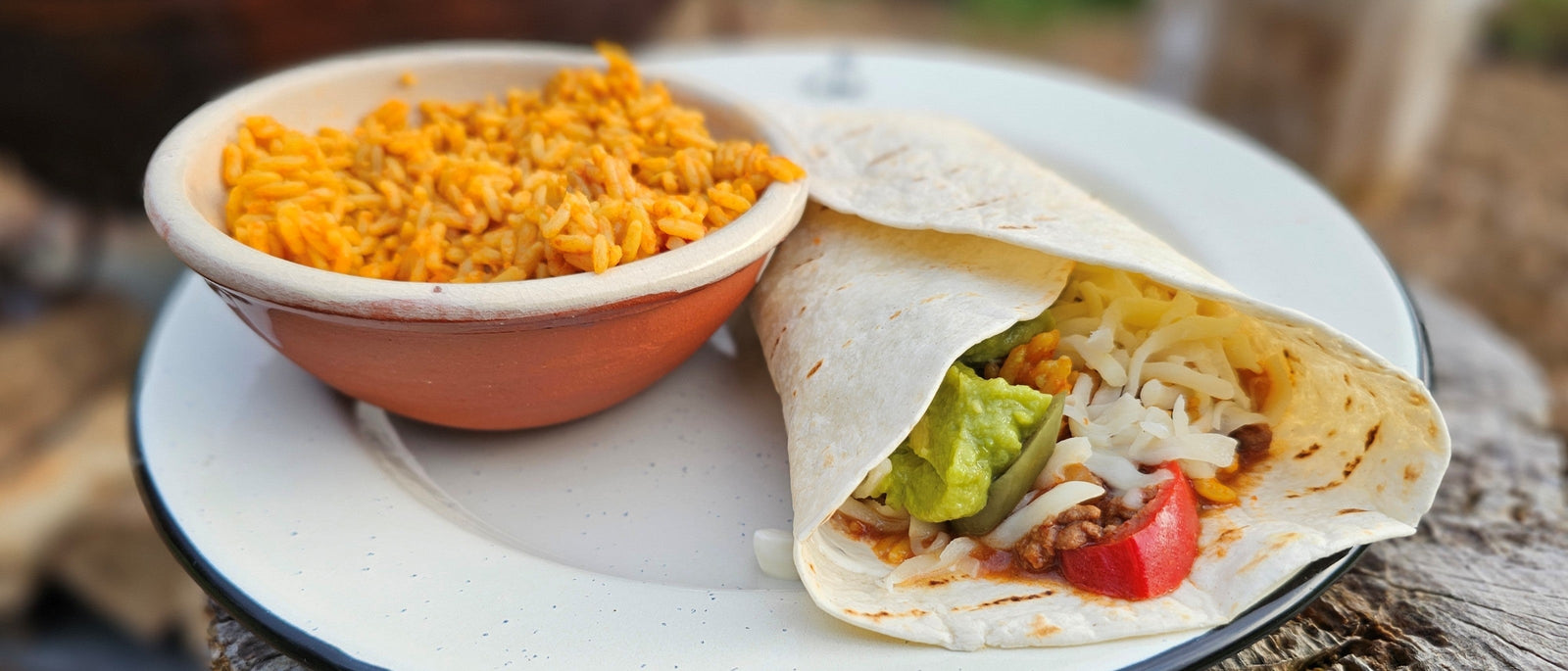 A folded campfire burrito filled with chilli, guacamole, cheese and tomato, served with a bowl of Spanish rice on an enamel camp plate