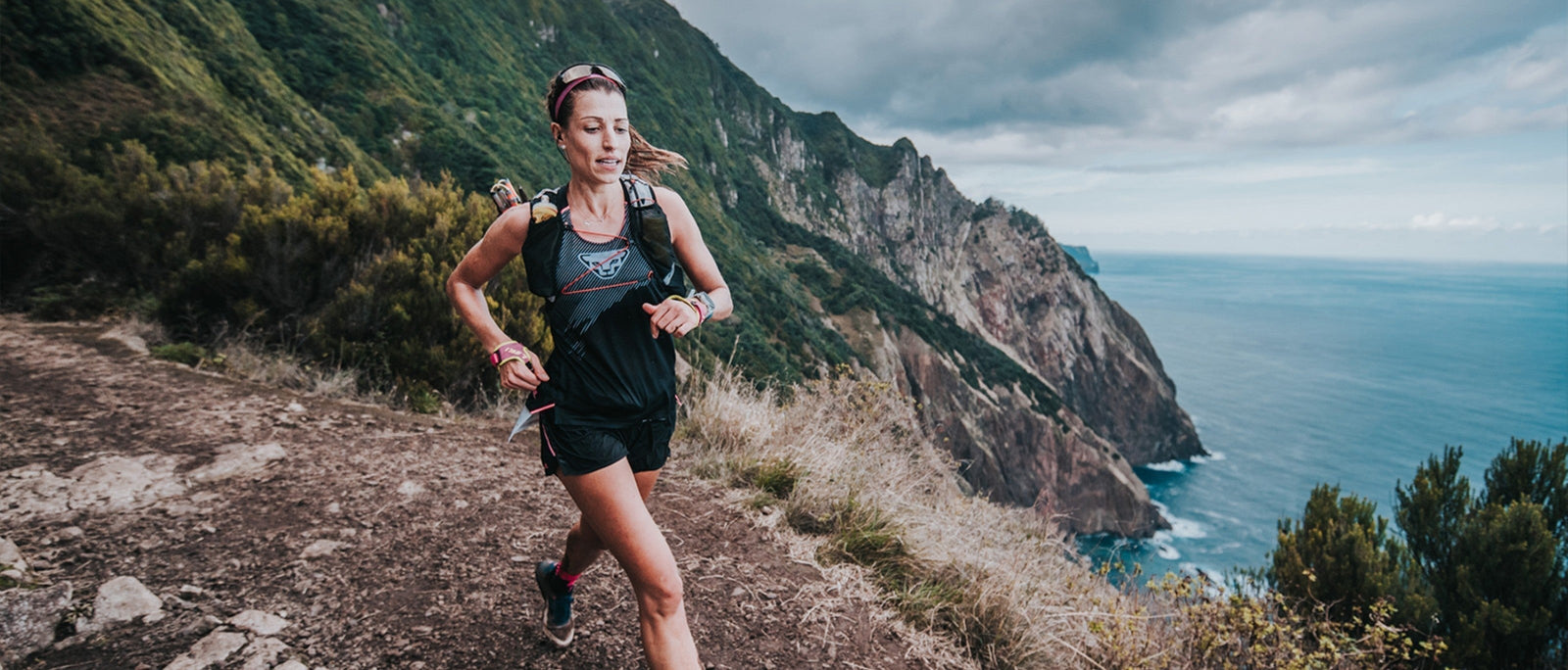 Female competitor in the Madeira Ultra Trail Race running along a rocky coastal path with a backdrop of steep cliffs plunging into deep blue sea. 
