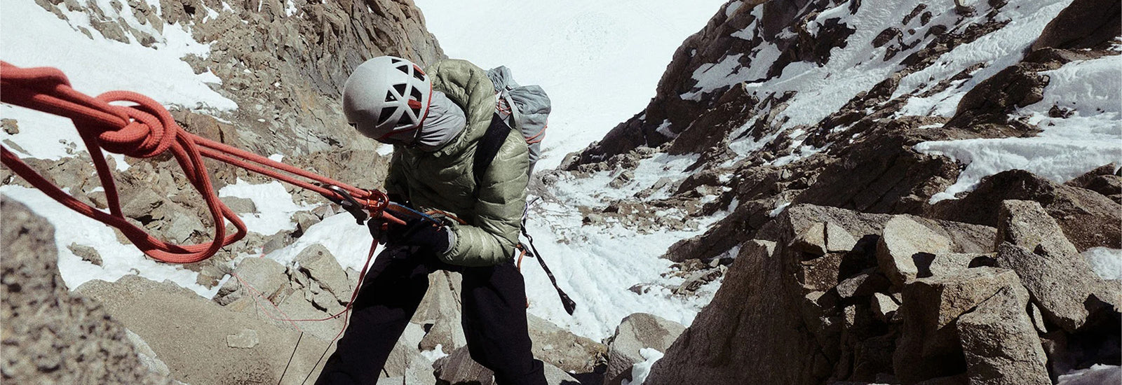 Climber in olive jacket and white helmet ascending snowy mountain with red rope and harness