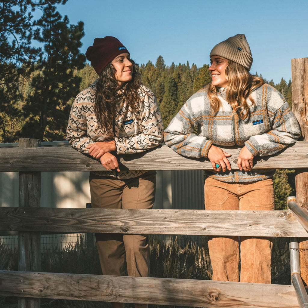 Two women wearing patterned fleece jackets and beanies lean on a wooden fence with coniferous forest behind them.
