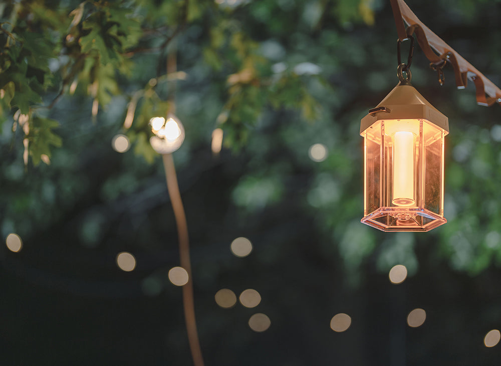 Illuminated copper camping lantern hanging from a leather strap among tree branches at dusk.