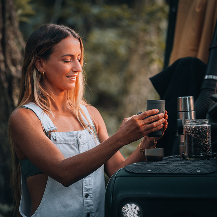Woman pouring hot water from a camping kettle into a metal cup at an outdoor camp stove in a woodland setting.