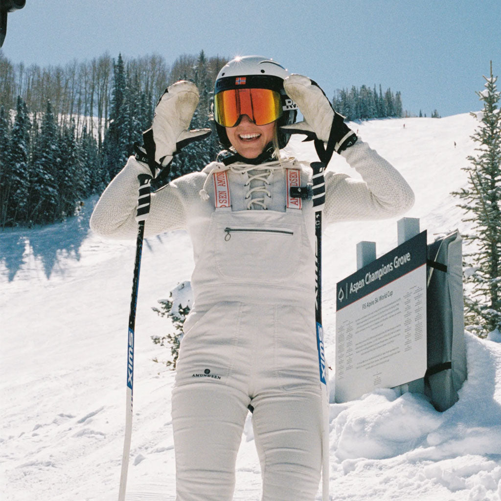 Skier in white Amundsen Sports suit and orange-tinted goggles at Aspen Champions Grove ski run.