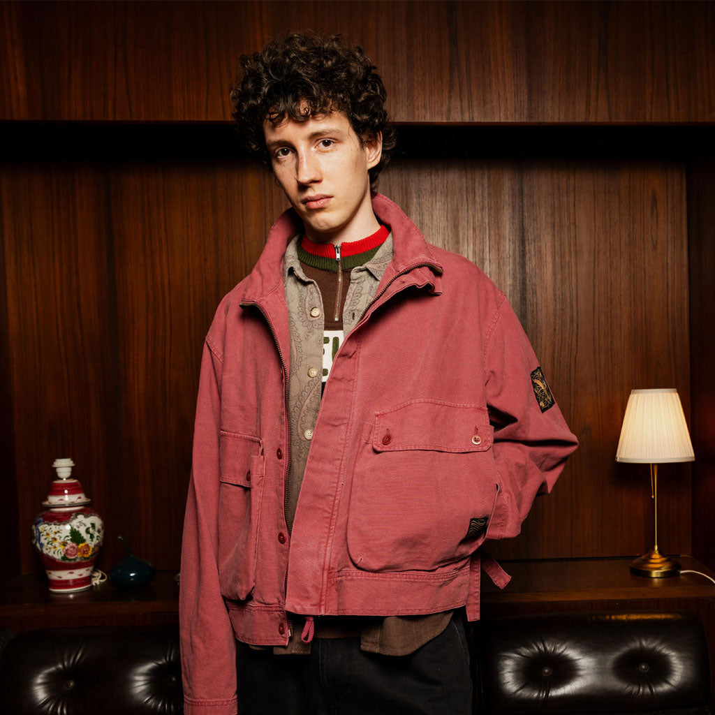 Person wearing a dusty rose denim jacket over a grey cardigan and red-striped collar, posed indoors against wooden panelling.