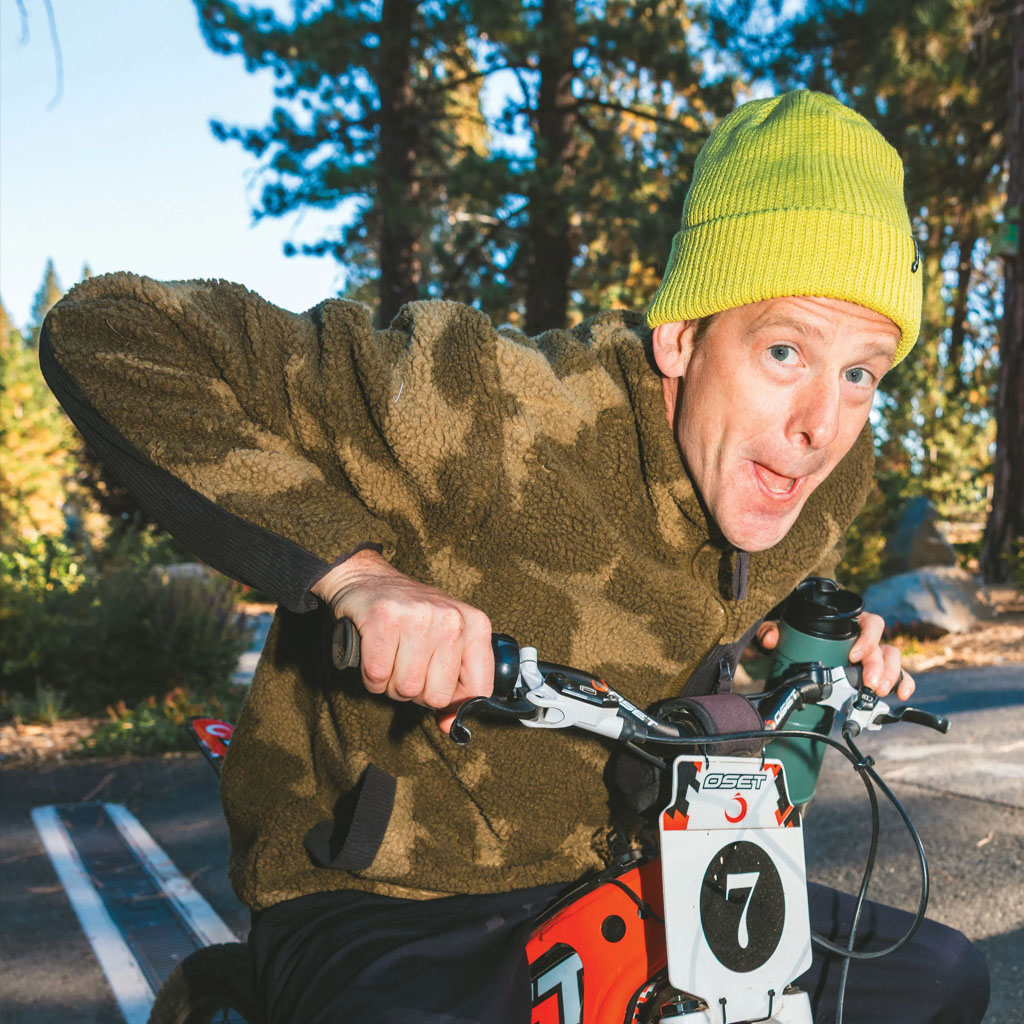 A person in a yellow beanie and olive Kavu jacket riding a mountain bike in a forest setting.