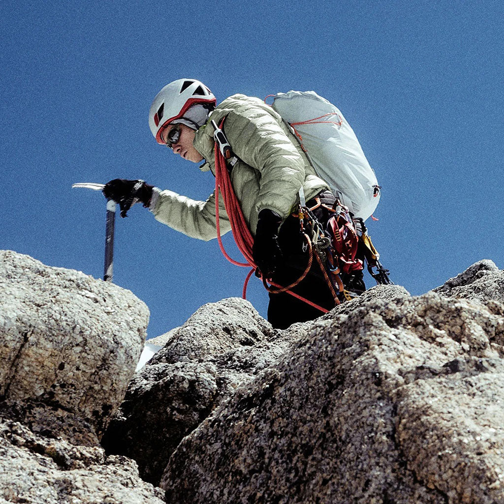 Rock climber in beige jacket and white helmet climbing rocky terrain against a clear blue sky.