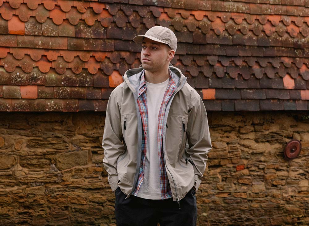 Man wearing beige jacket and cap against a brick wall, styled with Snow Peak outdoor gear.