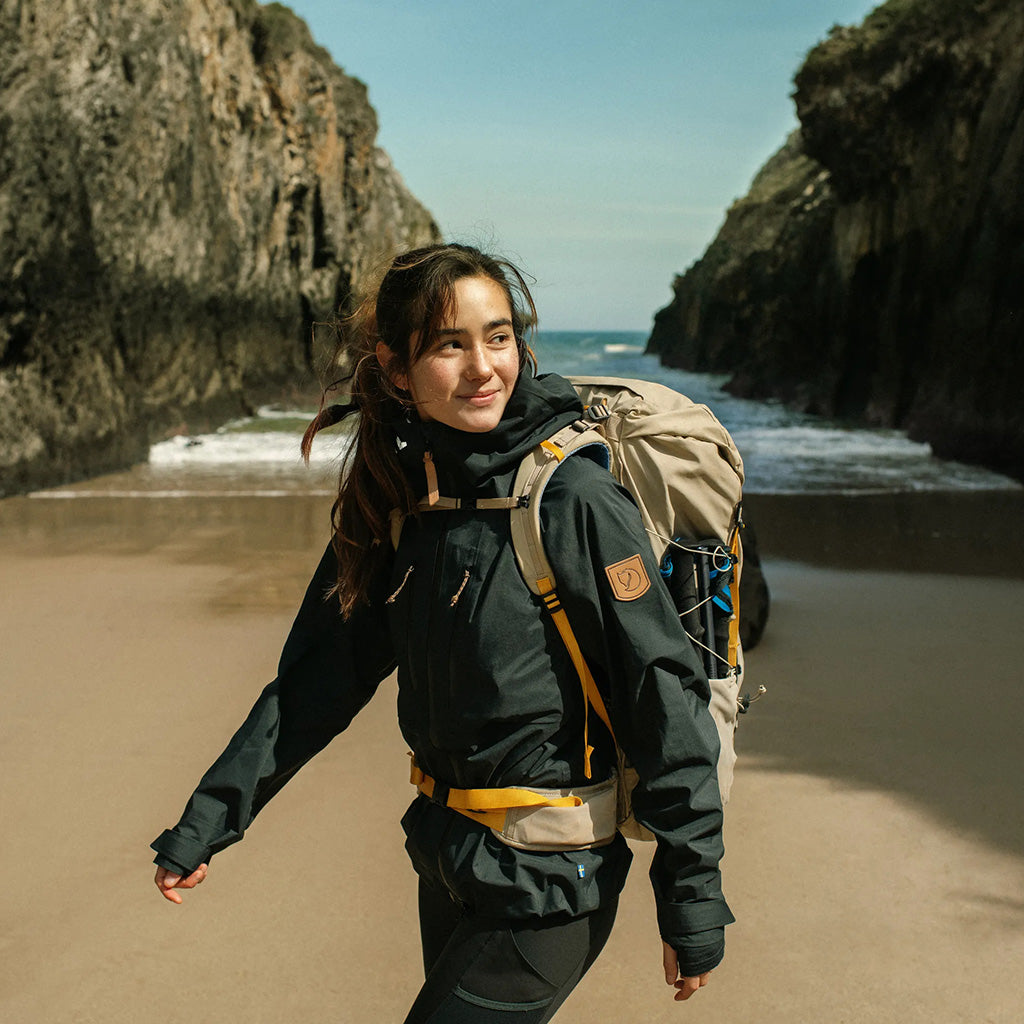 A hiker wearing a black jacket and backpack on a sandy beach between towering rock cliffs and the sea.